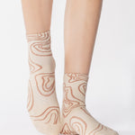 Woman wearing an off white ankle grip sock with flowing brown lines, made for pilates, barre, yoga, and beyond. 