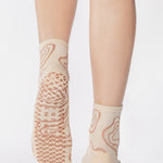 Woman wearing an off white ankle grip sock with flowing brown lines, made for pilates, barre, yoga, and beyond. 