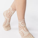 Woman wearing an off white ankle grip sock with flowing brown lines, made for pilates, barre, yoga, and beyond. 