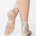 Woman wearing an off white ankle grip sock with flowing brown lines, made for pilates, barre, yoga, and beyond. 