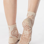 Woman wearing an off white ankle grip sock with flowing brown lines, made for pilates, barre, yoga, and beyond. 