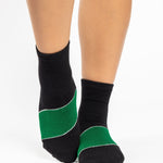 Woman wearing a black ankle grip sock with a solid green stripe in the center, made for pilates, barre, yoga, and beyond.  