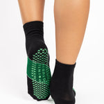 Woman wearing a black ankle grip sock with a solid green stripe in the center, made for pilates, barre, yoga, and beyond.  