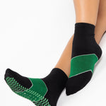 Woman wearing a black ankle grip sock with a solid green stripe in the center, made for pilates, barre, yoga, and beyond.  