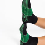 Woman wearing a black ankle grip sock with a solid green stripe in the center, made for pilates, barre, yoga, and beyond.  