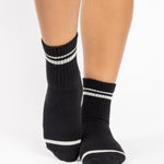 Women wearing a black ankle grip sock with white stripes on the toes and top ankle cuff made for Pilates, Barre, Yoga, and beyond. 