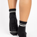 Women wearing a black ankle grip sock with white stripes on the toes and top ankle cuff made for Pilates, Barre, Yoga, and beyond. 