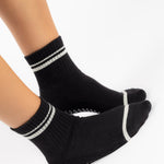 Women wearing a black ankle grip sock with white stripes on the toes and top ankle cuff made for Pilates, Barre, Yoga, and beyond. 