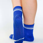 Women wearing a bright blue grip sock with white stripes on the toes and top ankle cuff made for Pilates, Barre, Yoga, and beyond. 