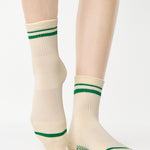 Women wearing a beige ankle grip sock with green stripes on the toes and top ankle cuff made for Pilates, Barre, Yoga, and beyond. 