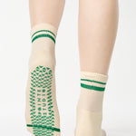 Women wearing a beige ankle grip sock with green stripes on the toes and top ankle cuff made for Pilates, Barre, Yoga, and beyond. 