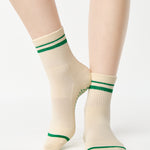 Women wearing a beige ankle grip sock with green stripes on the toes and top ankle cuff made for Pilates, Barre, Yoga, and beyond. 