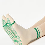 Women wearing a beige ankle grip sock with green stripes on the toes and top ankle cuff made for Pilates, Barre, Yoga, and beyond. 