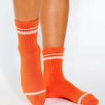 Women wearing a bright orange grip sock with white stripes on the toes and top ankle cuff made for Pilates, Barre, Yoga, and beyond. 