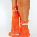 Women wearing a bright orange grip sock with white stripes on the toes and top ankle cuff made for Pilates, Barre, Yoga, and beyond. 