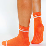 Women wearing a bright orange grip sock with white stripes on the toes and top ankle cuff made for Pilates, Barre, Yoga, and beyond. 