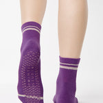 Women wearing a purple ankle grip sock with off white stripes on the toes and top ankle cuff made for Pilates, Barre, Yoga, and beyond. 