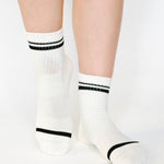 Women wearing a white ankle grip sock with black stripes on the toes and top ankle cuff made for Pilates, Barre, Yoga, and beyond. 