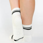 Women wearing a white ankle grip sock with black stripes on the toes and top ankle cuff made for Pilates, Barre, Yoga, and beyond. 