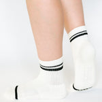 Women wearing a white ankle grip sock with black stripes on the toes and top ankle cuff made for Pilates, Barre, Yoga, and beyond. 
