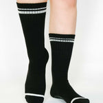 Women wearing a solid black crew grip sock with white stripes on the toes and top ankle cuff made for Pilates, Barre, Yoga, and beyond. 