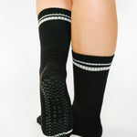 Women wearing a solid black crew grip sock with white stripes on the toes and top ankle cuff made for Pilates, Barre, Yoga, and beyond. 