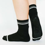 Women wearing a solid black crew grip sock with white stripes on the toes and top ankle cuff made for Pilates, Barre, Yoga, and beyond. 