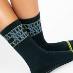 Woman wearing a black crew grip sock with "Sanity Not Vanity" written on the top cuff area in blue, made for pilates, barre, yoga, and beyond.