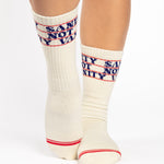 Woman wearing an off white crew grip sock with "Sanity Not Vanity" written on the top cuff area in blue, made for pilates, barre, yoga, and beyond.