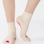 Women wearing a off white ankle grip sock with a red heart motif on the top made for Pilates, Barre, Yoga, and beyond. 