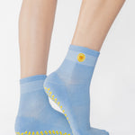 Woman wearing a light blue ankle grip sock with a yellow sun motif embroidered on the ankle side, made for pilates, barre, yoga, and beyond. 