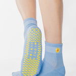 Woman wearing a light blue ankle grip sock with a yellow sun motif embroidered on the ankle side, made for pilates, barre, yoga, and beyond. 