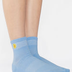 Woman wearing a light blue ankle grip sock with a yellow sun motif embroidered on the ankle side, made for pilates, barre, yoga, and beyond. 
