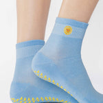Woman wearing a light blue ankle grip sock with a yellow sun motif embroidered on the ankle side, made for pilates, barre, yoga, and beyond. 