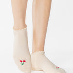 Women wearing a solid off white ivory full foot grip sock with a ruffle cuff and with a red cherry on the top of the toes, made for pilates, barre, yoga, and beyond.