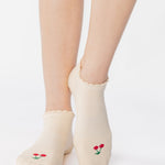 Women wearing a solid off white ivory full foot grip sock with a ruffle cuff and with a red cherry on the top of the toes, made for pilates, barre, yoga, and beyond.