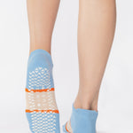 Woman wearing a light blue full foot grip sock with a bold off white stripe with a contrasting orange outline, made for pilates, barre, yoga, and beyond. 