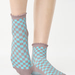 Woman wearing a checkered light blue with gray ankle grip sock with a ruffled cuff made for Pilates, Barre, Yoga, and beyond. 