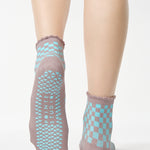 Woman wearing a checkered light blue with gray ankle grip sock with a ruffled cuff made for Pilates, Barre, Yoga, and beyond. 