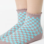 Woman wearing a checkered light blue with gray ankle grip sock with a ruffled cuff made for Pilates, Barre, Yoga, and beyond. 