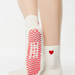 Women wearing a solid white ankle grip sock with a red heart on the ankle made for Pilates, Barre, Yoga, and beyond. 