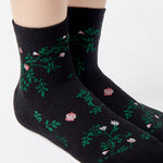 Woman wearing a black ankle grip sock with green rose vines and pink flowers made for Pilates, Barre, Yoga, and beyond.