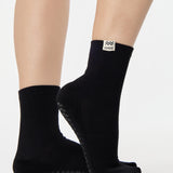 Woman wearing a solid black ankle grip sock with aquarius zodiac sign embroidered on the side. Made for pilates, barre, yoga, and beyond. 