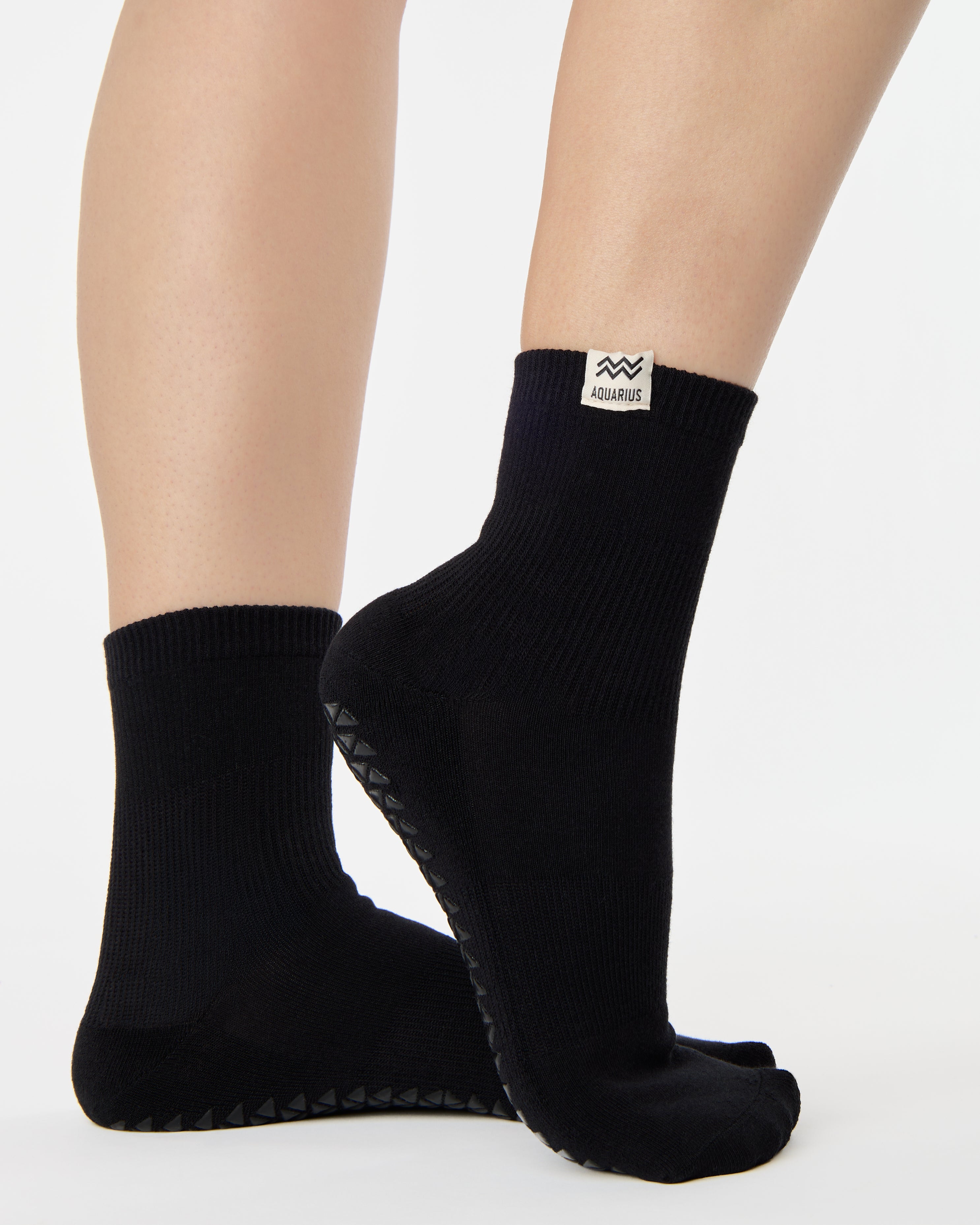 Woman wearing a solid black ankle grip sock with aquarius zodiac sign embroidered on the side. Made for pilates, barre, yoga, and beyond. 