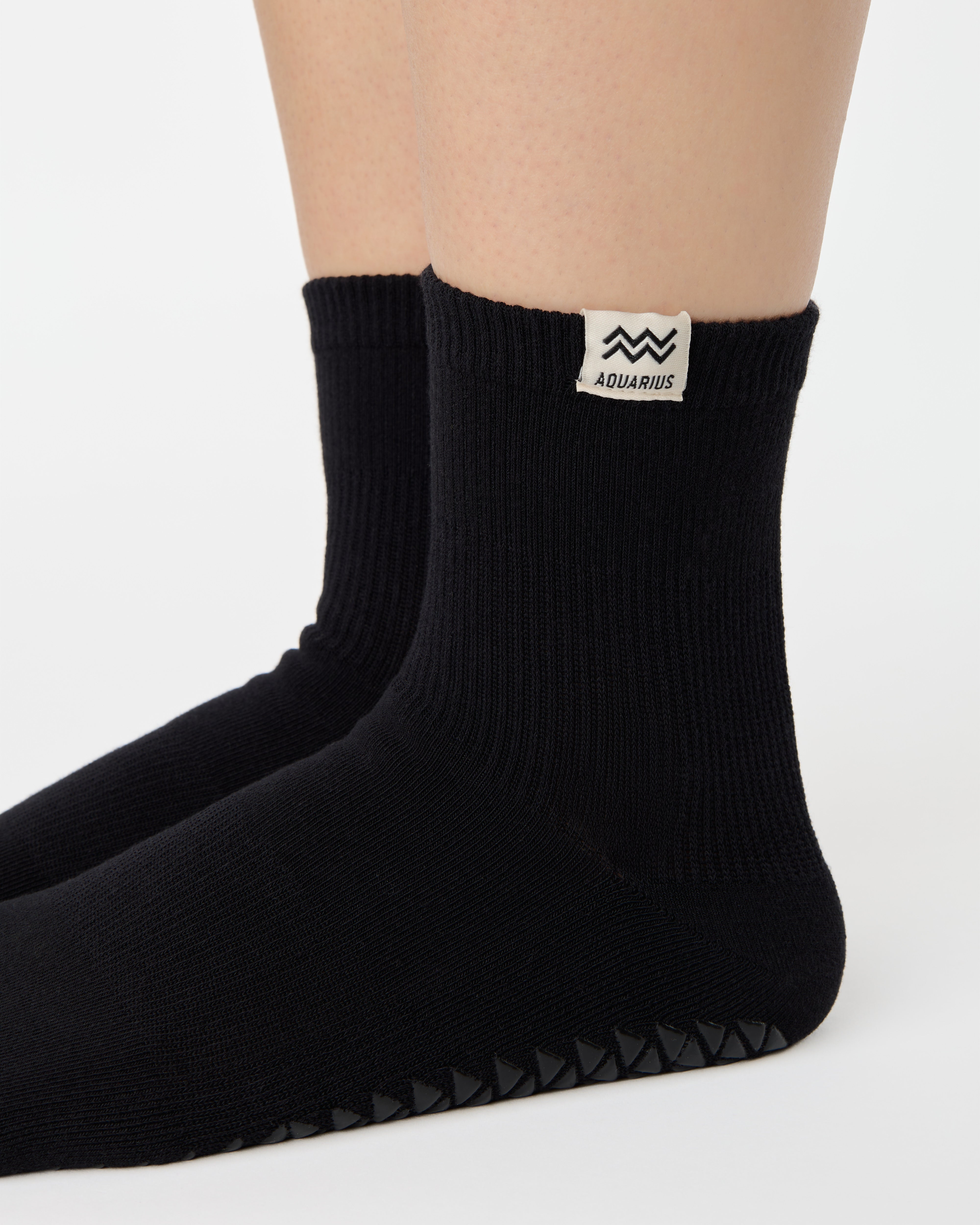 Woman wearing a solid black ankle grip sock with aquarius zodiac sign embroidered on the side. Made for pilates, barre, yoga, and beyond. 