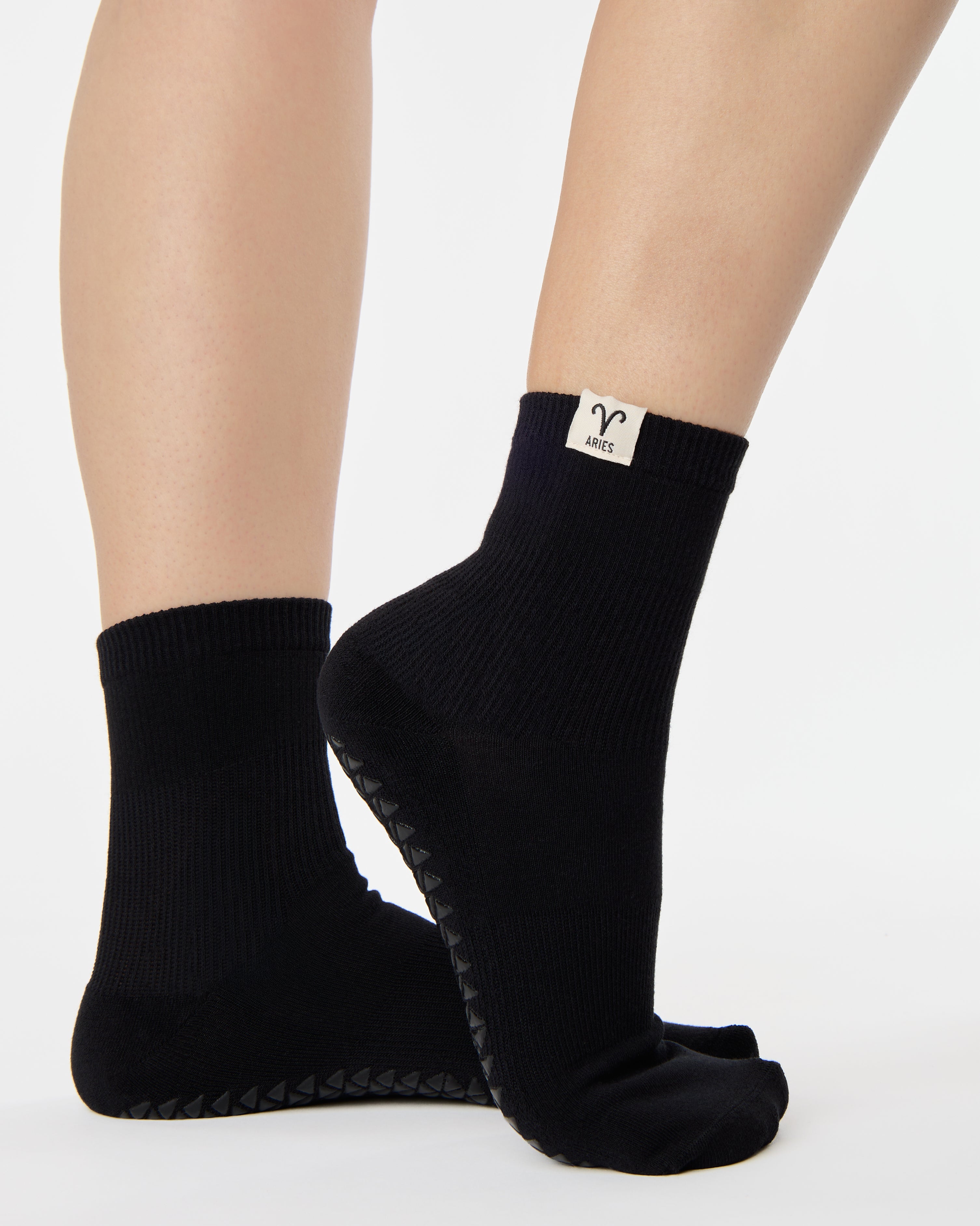 Woman wearing a solid black ankle grip sock with aries zodiac sign embroidered on the side. Made for pilates, barre, yoga, and beyond. 