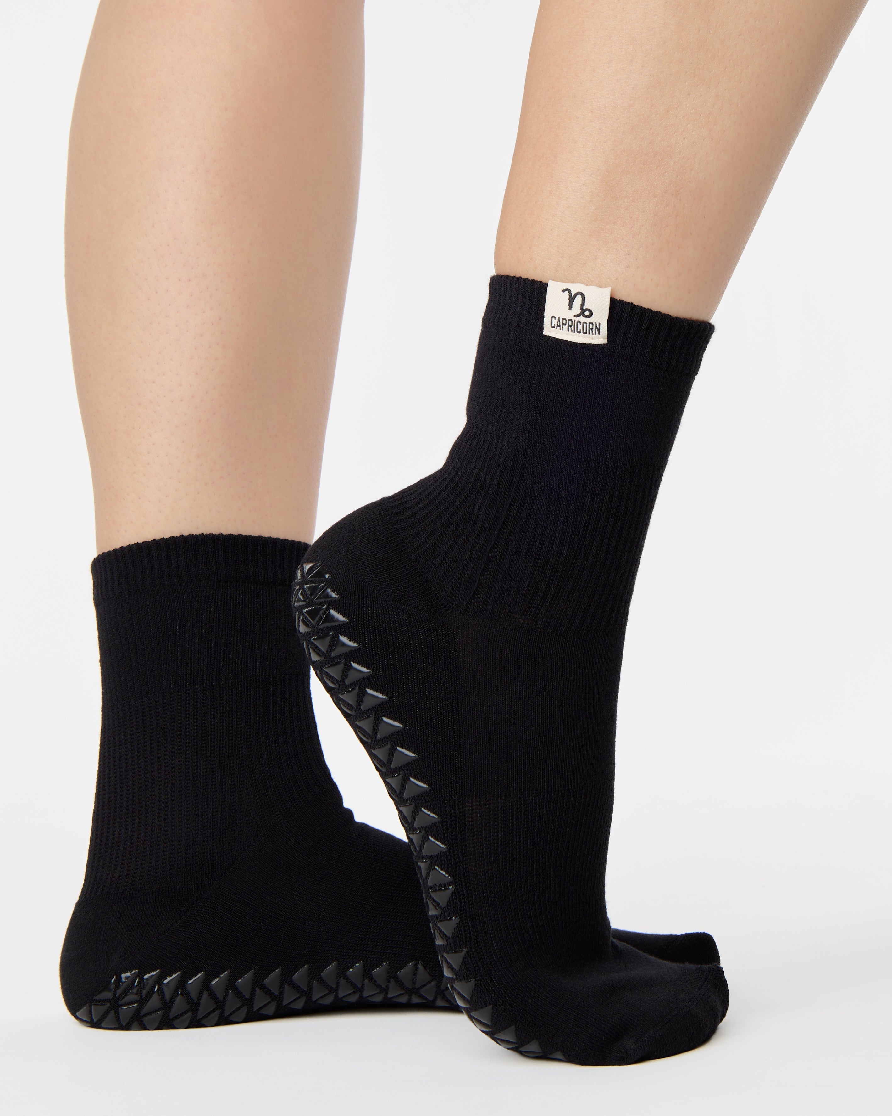Woman wearing a solid black ankle grip sock with capricorn zodiac sign embroidered on the side. Made for pilates, barre, yoga, and beyond. 