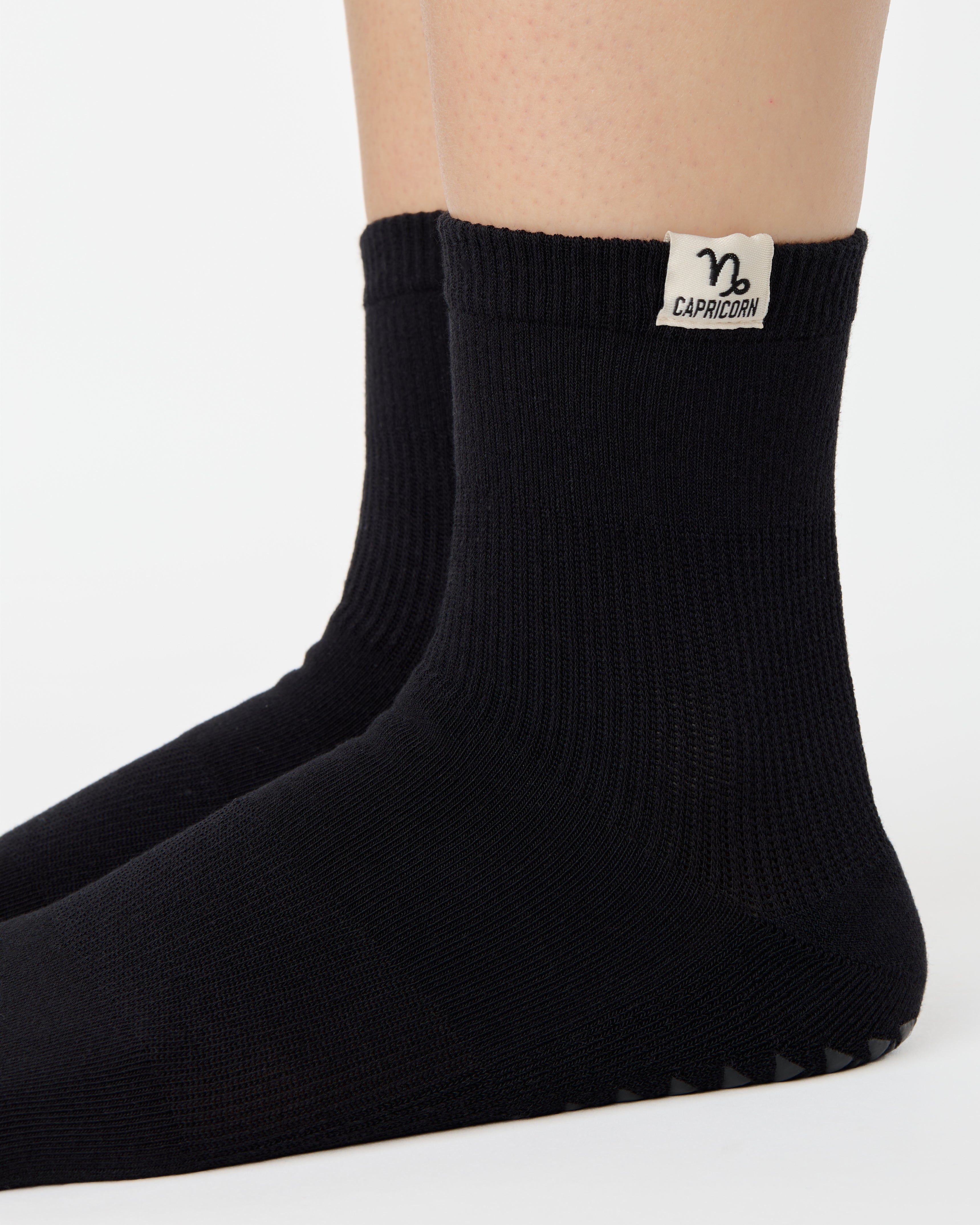 Woman wearing a solid black ankle grip sock with capricorn zodiac sign embroidered on the side. Made for pilates, barre, yoga, and beyond. 