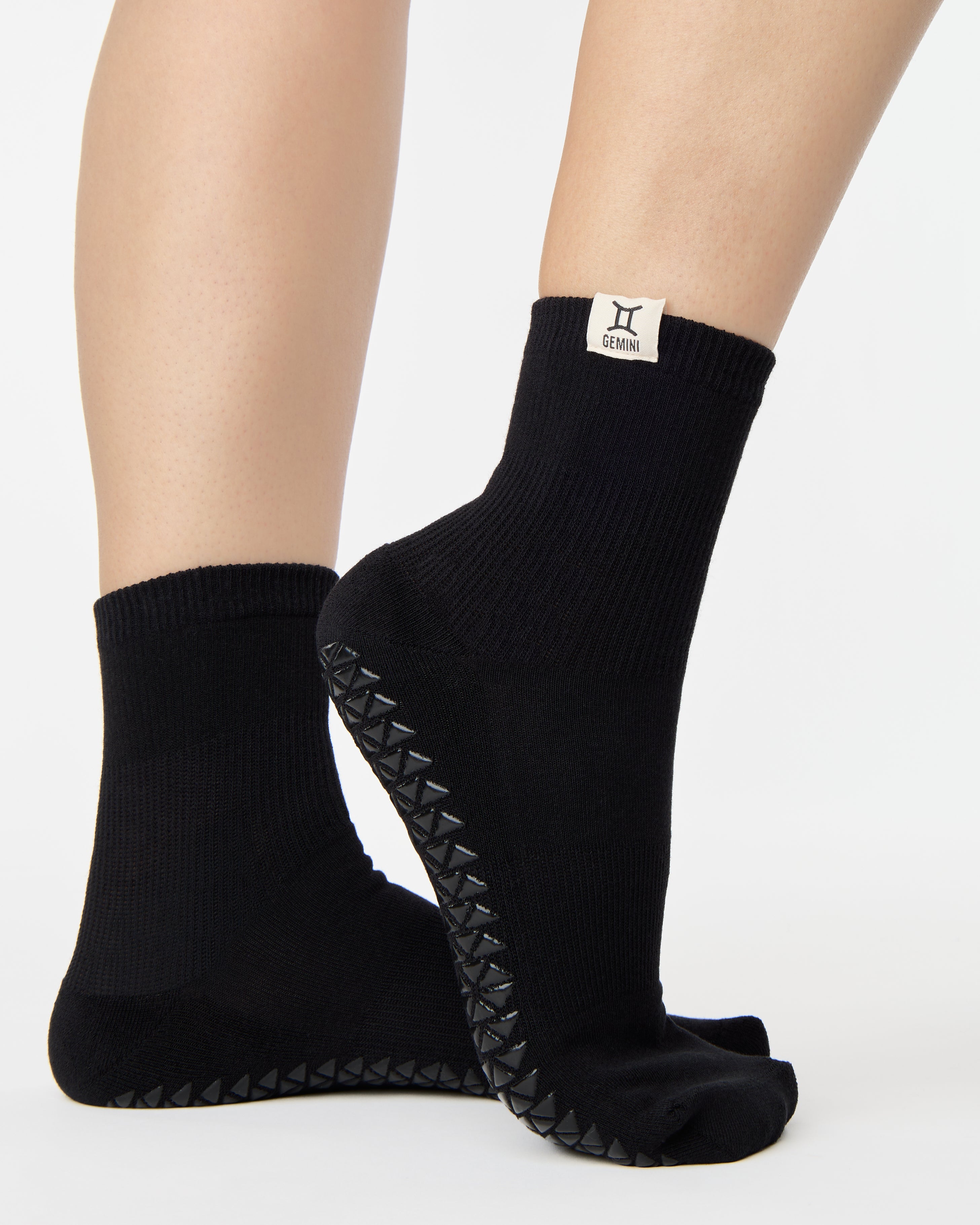 Woman wearing a solid black ankle grip sock with gemini zodiac sign embroidered on the side. Made for pilates, barre, yoga, and beyond. 