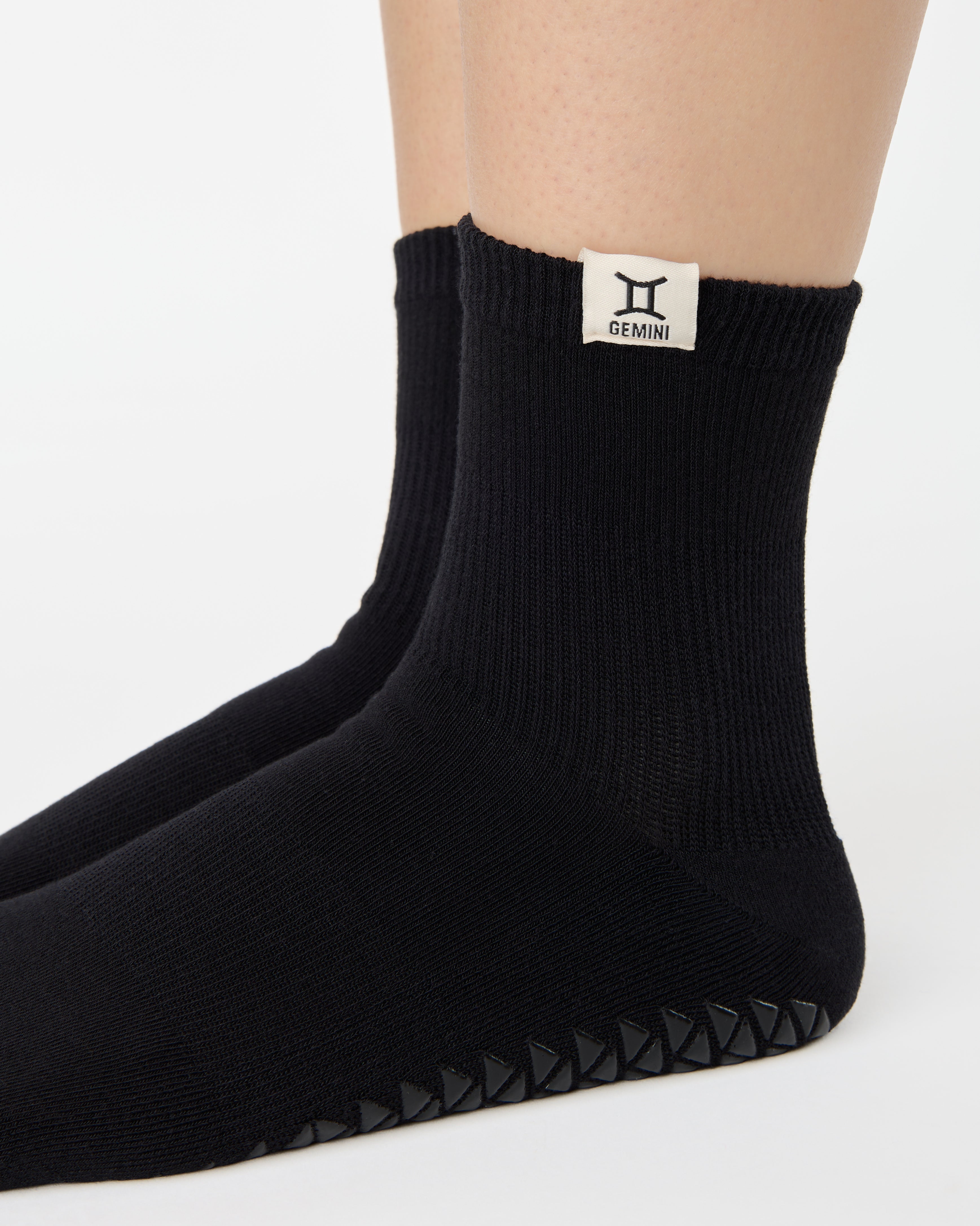 Woman wearing a solid black ankle grip sock with gemini zodiac sign embroidered on the side. Made for pilates, barre, yoga, and beyond. 
